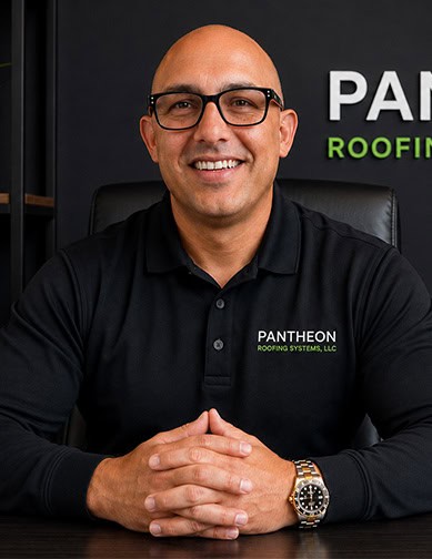 A man wearing glasses and a black "Pantheon Roofing Systems, LLC" shirt sits at a desk, smiling, with his hands folded in front of him.