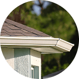 Close-up view of a house roof corner with asphalt shingles, white soffit, fascia, and rain gutter installed by Pantheon Roofing in Florida, set against a blurred green background.
