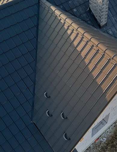 A close-up view of a dark metal roof by Pantheon Roofing with visible ridges, vent pipes, and a brick chimney on a residential Florida building.