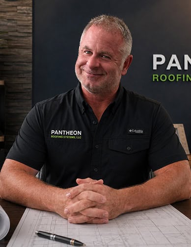 A man in a black Pantheon Roofing Systems shirt sits at a desk with architectural plans and a pen, smiling at the camera.